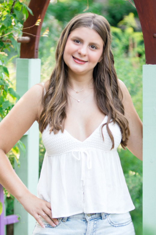 A young woman with long brown hair stands outdoors, smiling, under a wooden garden archway with green vines—perfect for senior pictures in Detroit with flowers. She wears a white sleeveless top and blue jeans amid greenery and purple fence accents.