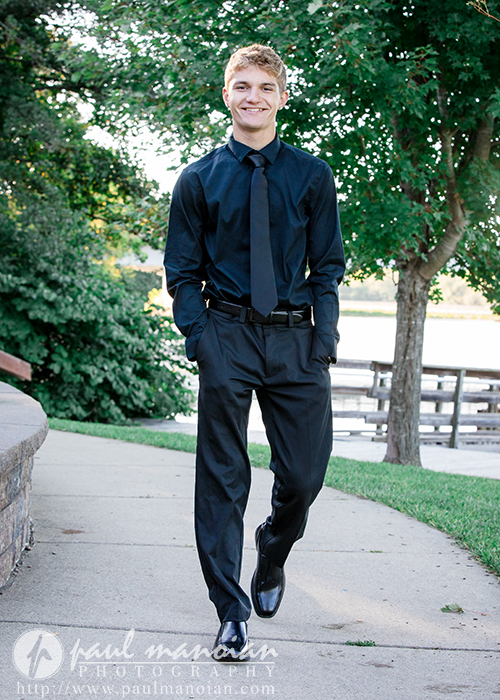 A young man wearing a black dress shirt, black tie, and black pants walks on a path near a lake, smiling during his Ann Arbor senior pictures session. Green trees and a wooden dock are visible in the background.