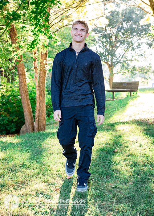 A young man in a dark long-sleeve shirt and cargo pants walks outside on a grassy area, smiling during his Ann Arbor senior pictures session. Sunlight filters through the trees behind him, with a wooden bench visible in the background.