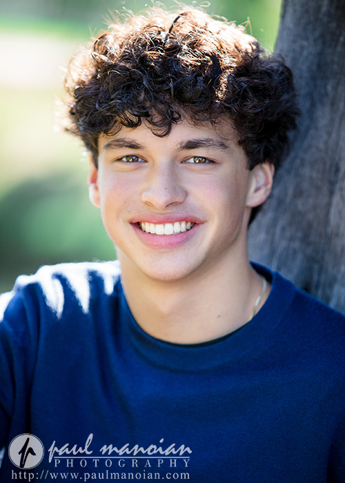 A young man with curly brown hair and a blue shirt smiles at the camera during his Clinton Township senior pictures session, sunlight highlighting his face as he sits outdoors.