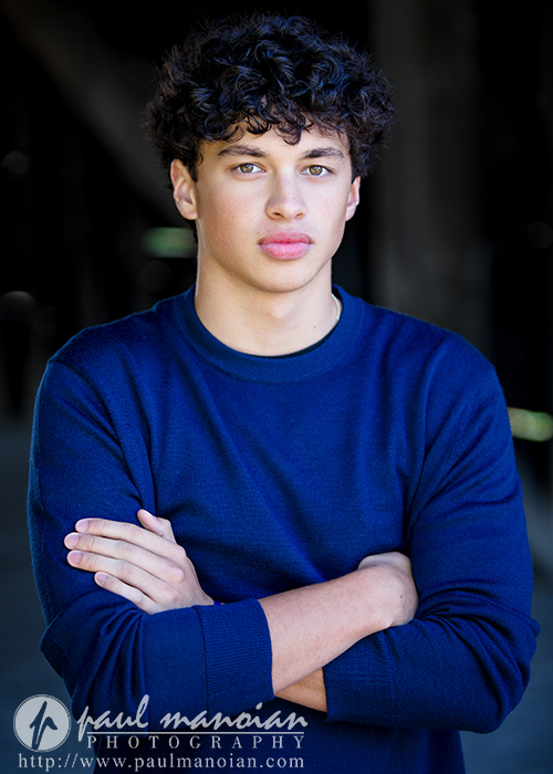 A young man with curly dark hair, wearing a blue sweater, stands with arms crossed and looks at the camera. Captured during a Clinton Township senior pictures session, the dark, blurred background adds focus. Watermarked by Paul Manoian Photography.