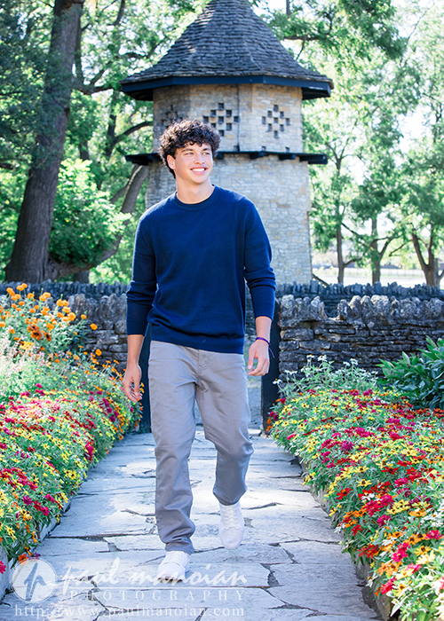 A young man with curly hair, wearing a navy sweater and gray pants, walks along a stone path lined with colorful flowers during his Clinton Township senior pictures session, with a stone building and green trees in the background.