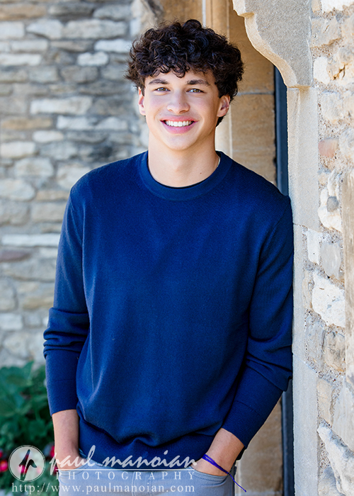 A young person with curly brown hair wearing a blue sweater stands and smiles in front of a stone wall, posing with one hand in their pocket during their Clinton Township senior pictures session.