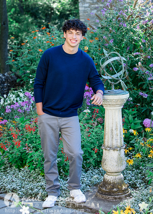A young man with curly dark hair, wearing a navy sweater and gray pants, stands next to a decorative stone pedestal in a colorful garden full of flowers, smiling at the camera during his Clinton Township senior pictures session.