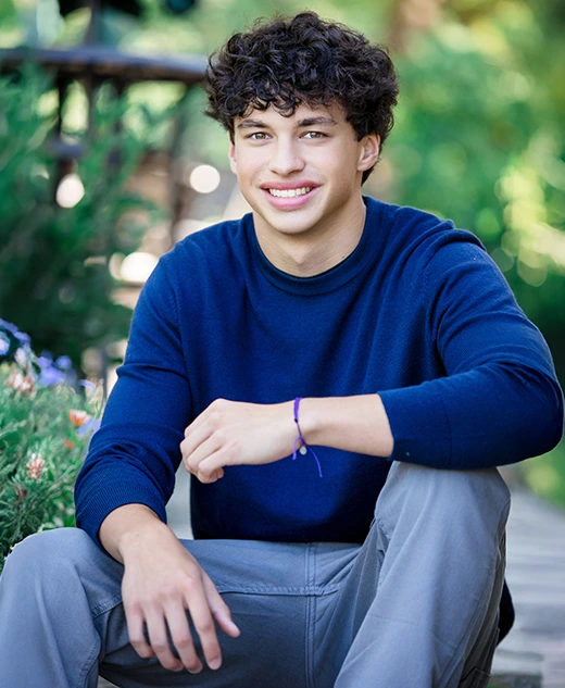 A high school senior boy with curly hair, wearing a blue sweater and gray pants, sits outdoors on a wooden path surrounded by greenery and flowers, smiling at the camera during his Clinton Township senior pictures session.
