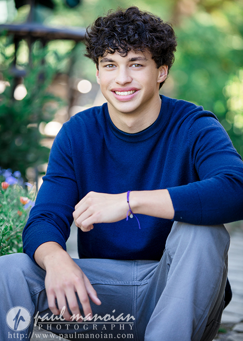 A young man with curly brown hair, wearing a blue sweater and gray pants, sits outdoors on a wooden pathway during his Clinton Township senior pictures session, smiling at the camera. Greenery is visible in the background.