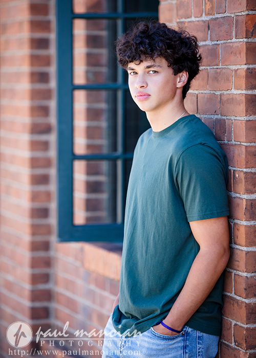 A young man with curly dark hair wearing a green t-shirt and jeans leans against a brick wall near a window, looking at the camera with a neutral expression during his Clinton Township senior pictures session.