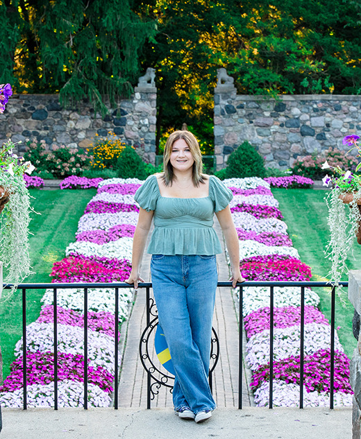 A high school senior in a green top and jeans stands smiling in front of colorful rows of pink and white flowers during a session for Livonia Senior Pictures at Cranbrook House & Gardens, flanked by two large flowerpots with a stone wall and trees behind her.