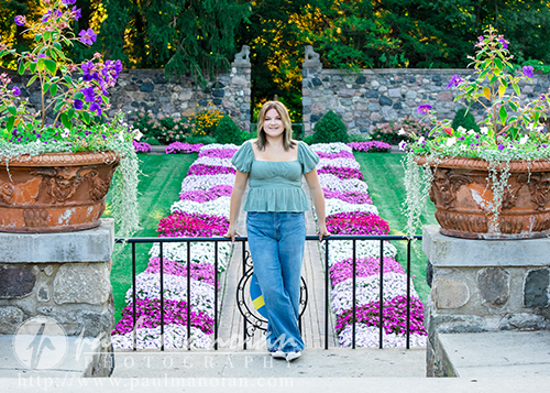 A high school senior girl stands smiling on a stone walkway between two large flower pots during a session for Livonia Senior Pictures at Cranbrook House & Gardens, with vibrant flower beds and a stone wall in the background on a sunny day.