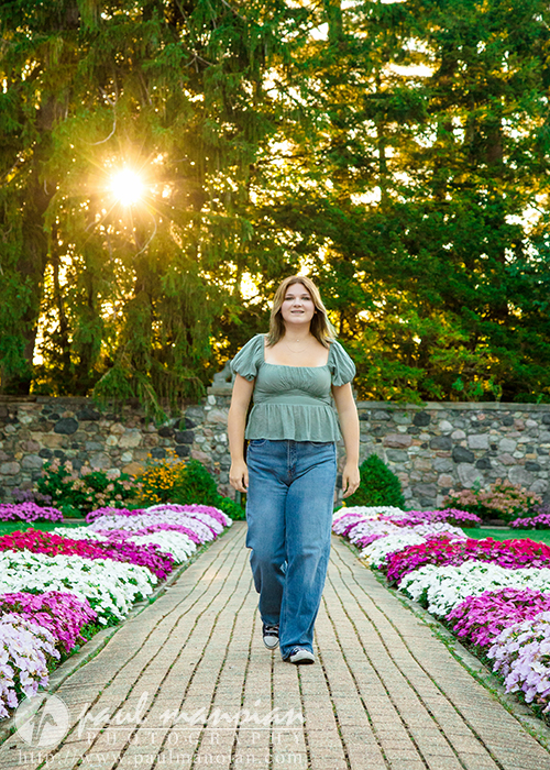 A high school senior girl in a green top and jeans walks on a brick path lined with colorful flowers during a session for Livonia Senior Pictures at Cranbrook House & Gardens, with sunlight streaming through tall trees and a stone wall in the background.
