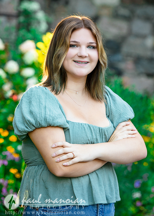 A high school senior girl with shoulder-length brown hair, wearing a light green blouse, stands outdoors with her arms crossed, smiling at the camera during a session for Livonia Senior Pictures at Cranbrook House & Gardens. Colorful flowers and greenery blur in the background.