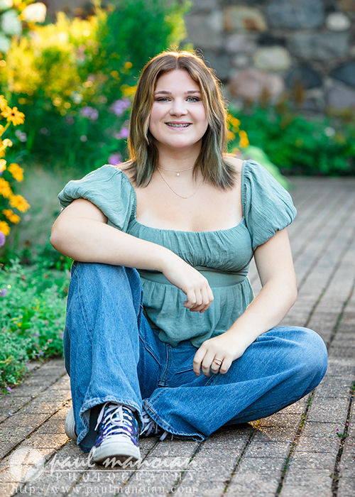 A high school senior girl with shoulder-length hair, wearing a green top and blue jeans, sits cross-legged on a brick path at Cranbrook House & Gardens for her Livonia Senior Pictures session, smiling at the camera amid bright flowers and lush greenery.