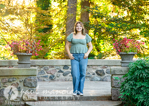 A high school senior girl in a green top and blue jeans stands smiling on a stone path between two large planters, with trees and greenery in the background during a sunny session for Livonia Senior Pictures at Cranbrook House & Gardens.