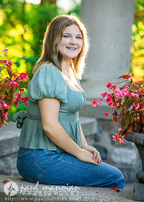 A high school senior girl with shoulder-length brown hair, wearing a green top and blue jeans, sits on a stone ledge beside pink flowers, smiling outdoors in bright, natural light during a session for Livonia Senior Pictures at Cranbrook House & Gardens.