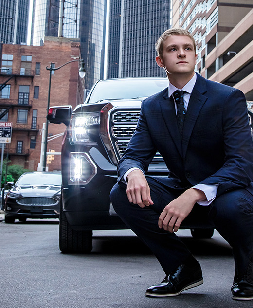 A young man in a navy blue suit squats confidently in front of a black SUV on a city street, with tall buildings and parked cars in the background.