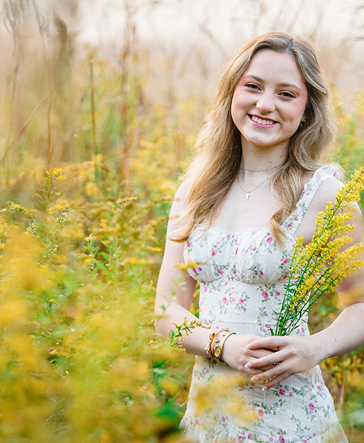 A young woman in a floral dress stands in a sunlit field of yellow wildflowers during her Novi senior pictures session, smiling and holding a small bouquet, with tall grass and plants surrounding her.