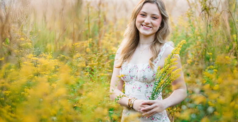 A young woman in a floral dress stands in a sunlit field of yellow wildflowers during her Novi senior pictures session, smiling and holding a small bouquet, with tall grass and plants surrounding her.