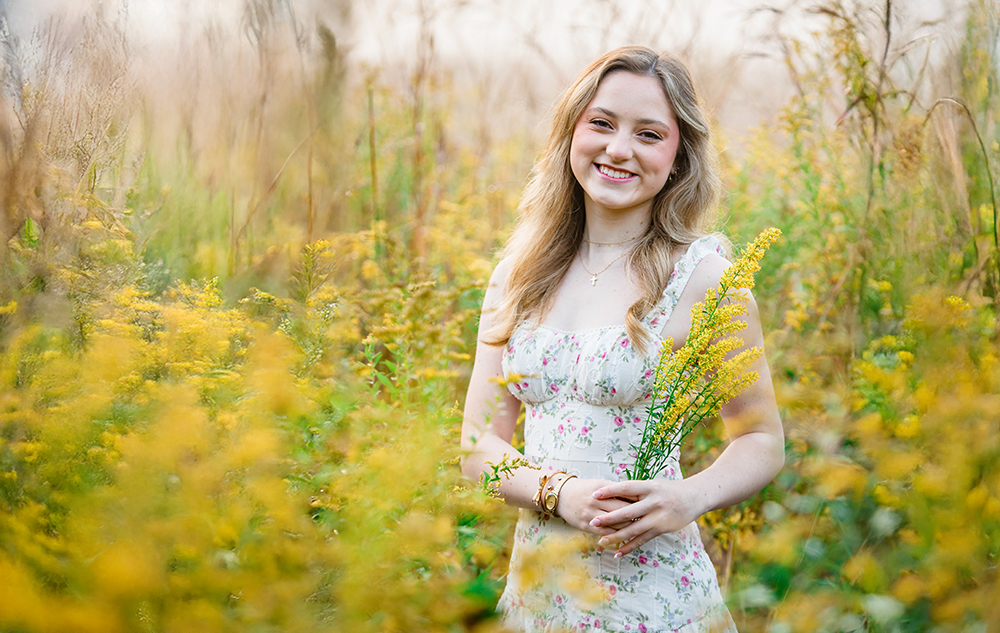 A young woman in a floral dress stands in a sunlit field of yellow wildflowers during her Novi senior pictures session, smiling and holding a small bouquet, with tall grass and plants surrounding her.