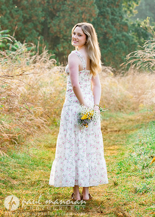 A high school senior girl in a long, floral dress stands on a grassy path during her Novi senior pictures session, holding a small bouquet of wildflowers behind her back as she looks over her shoulder, surrounded by tall grass and greenery.