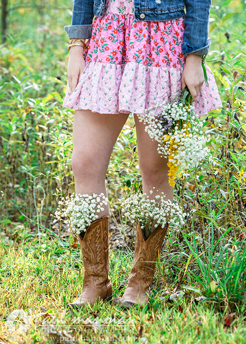 A person wearing a pink floral dress, denim jacket, and cowboy boots stands in grass during a Novi senior pictures session. Wildflowers are held in one hand and tucked into the boots, creating a whimsical, nature-inspired look.
