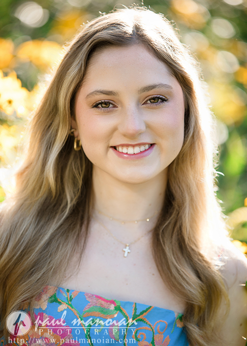 A young woman with long blonde hair smiles outdoors during her Novi senior pictures session, wearing a blue floral dress and a cross necklace. Sunlight and yellow flowers create a bright, cheerful background.