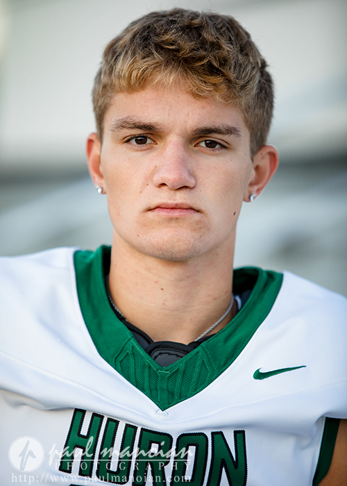 A young male athlete with short light brown hair and earrings wears a white and green "Hurons" jersey, looking directly at the camera with a serious expression during his football senior pictures session.