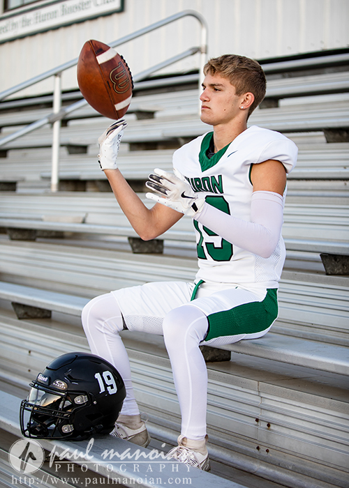 A young male football player in a white and green uniform sits on bleachers, tossing a football in one hand during his football senior pictures session. His helmet rests on the bench beside him, and he appears focused.