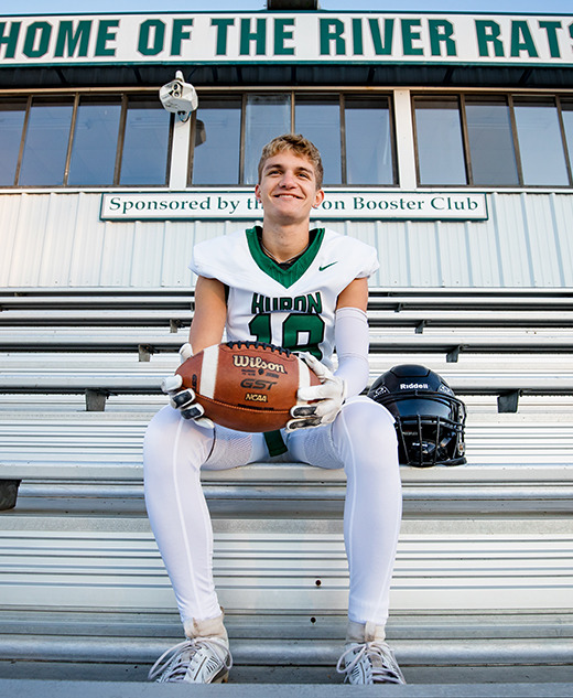 A football player in uniform sits on bleachers holding a football, with his helmet beside him. Behind him is a stadium sign that reads "Home of the River Rats." He smiles at the camera during his football senior pictures session.