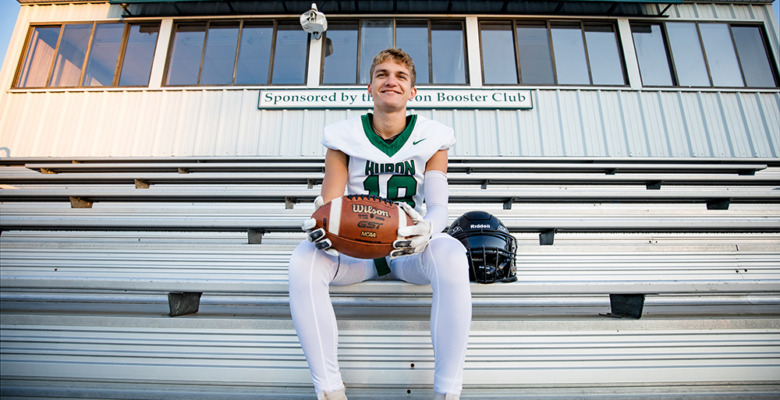 A football player in uniform sits on bleachers holding a football, with his helmet beside him. Behind him is a stadium sign that reads "Home of the River Rats." He smiles at the camera during his football senior pictures session.