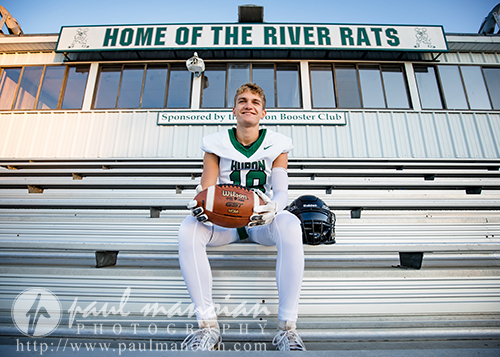 A young football player in uniform sits on bleachers holding a football, with a helmet beside him—capturing the spirit of a football senior pictures session. Behind him, a building displays a sign reading "Home of the River Rats.
