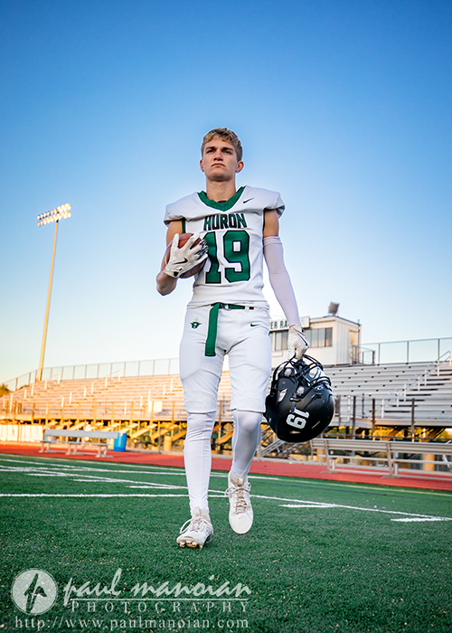A high school football player in a white Huron uniform, holding a football and black helmet, walks on the field during his football senior pictures session with empty bleachers behind him under a clear blue sky.