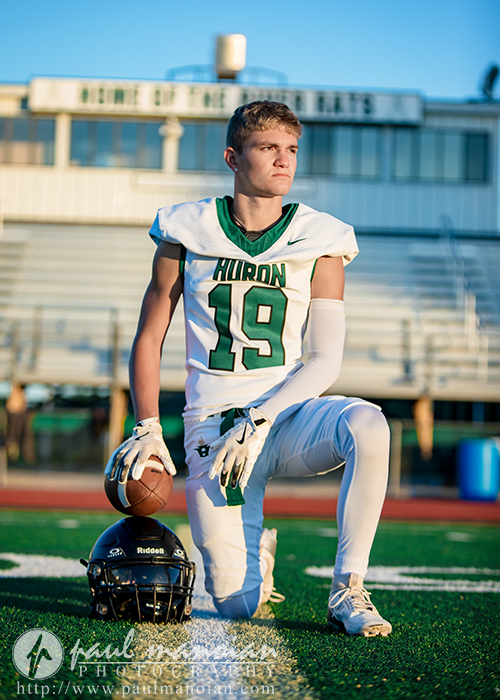 A high school football player in a white and green "Huron" uniform (#19) kneels on the field during his football senior pictures session, holding a football with his helmet beside him. Bleachers and a "Home of the River Rats" sign are visible behind him.