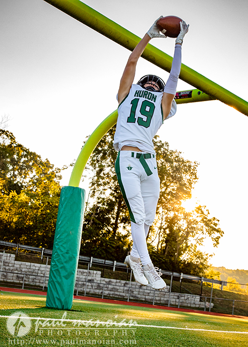 A football player in a white and green uniform, number 19, jumps and hangs from a goalpost while holding a football during a sunset football senior pictures session on an outdoor field.