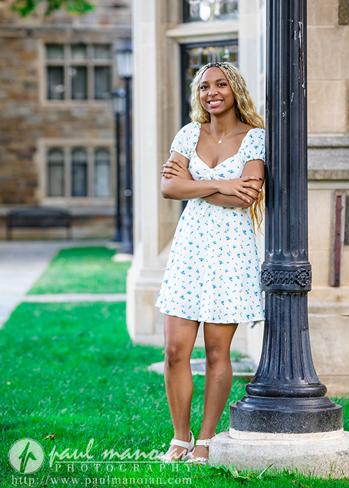 A high school senior in a light blue floral dress stands with arms crossed, leaning against a black lamppost on a grassy area near a stone building, smiling at the camera during her Livonia senior pictures session.