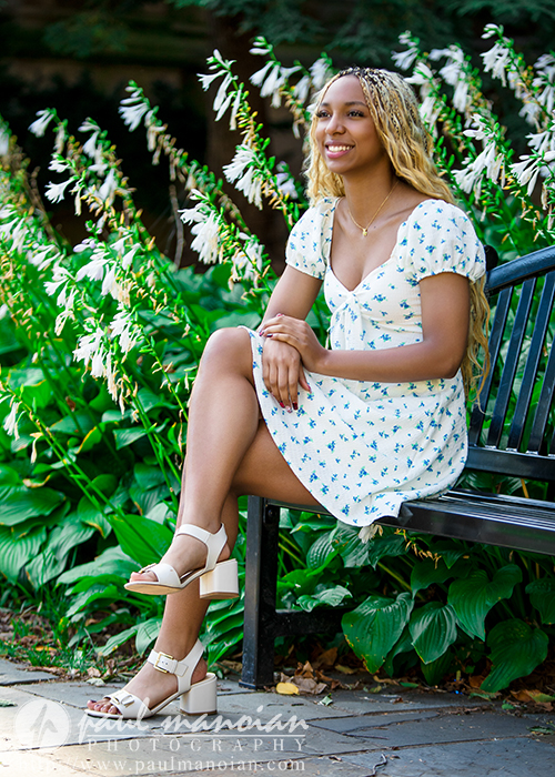 A high school senior in a white floral dress sits on a black bench, smiling with hands on her knee, surrounded by lush green plants and bright sunlight during a Livonia senior pictures session in a garden setting.