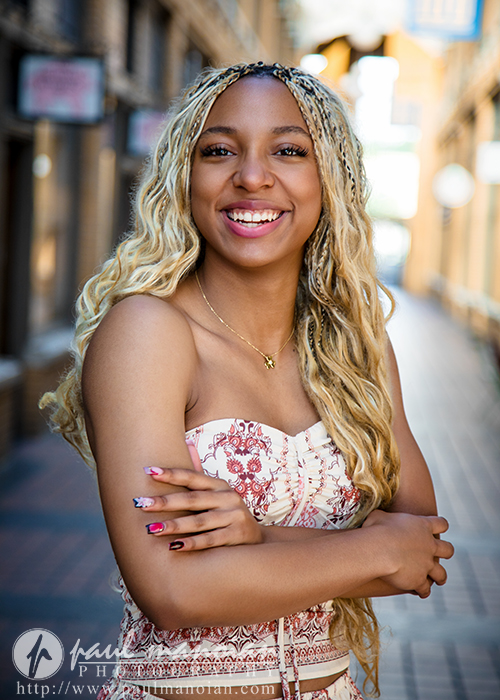 A high school senior with long blonde braids smiles brightly, standing outdoors in a sunlit, urban alleyway during her Livonia senior pictures session. She wears a strapless, patterned dress and has her arms crossed.
