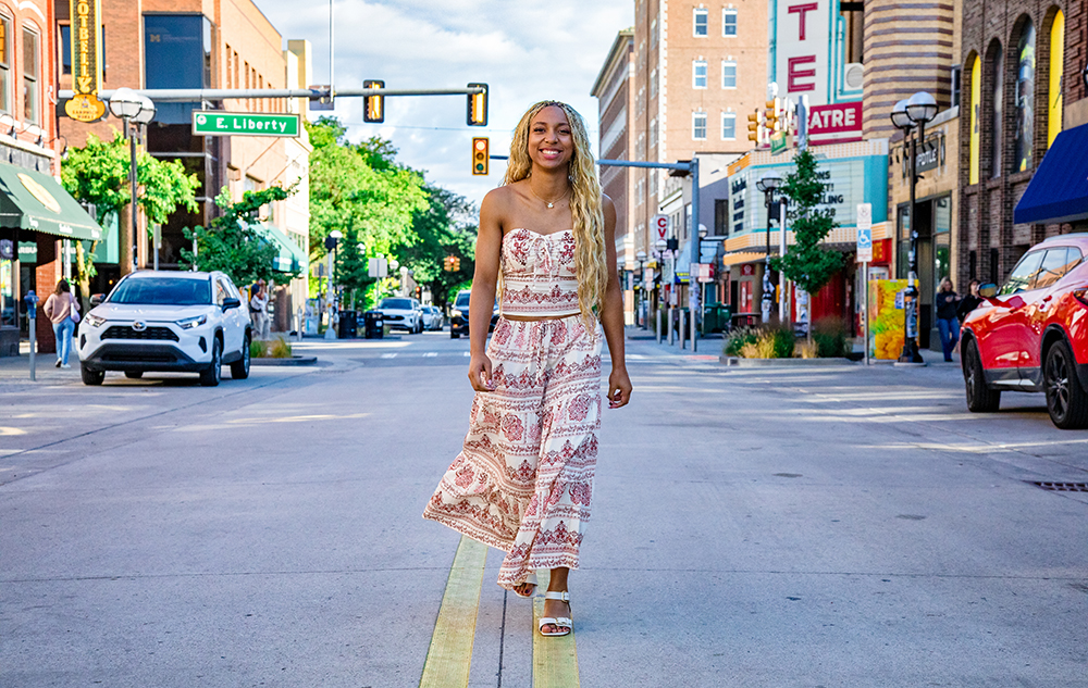 A woman with long blonde hair wearing a patterned strapless top and matching pants walks confidently down the middle of a city street lined with shops, cars, and trees on a sunny day, capturing the vibrant spirit of Livonia senior pictures.