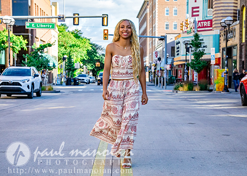 A high school senior in a patterned strapless jumpsuit walks confidently down a city street during her Livonia senior pictures session, with cars and buildings in the background on a sunny day.