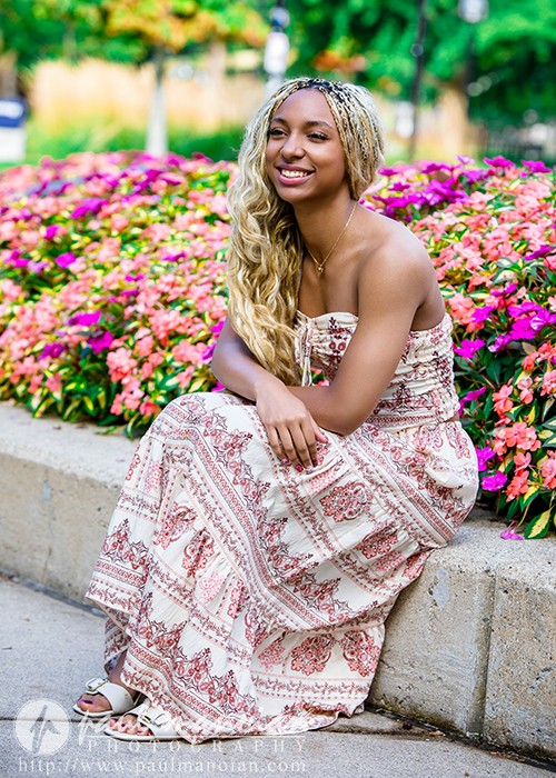 A high school senior with long blonde braids and a patterned headband sits on a concrete ledge in front of vibrant pink flowers during her Livonia senior pictures session, wearing a strapless, patterned maxi dress and sandals, smiling at the camera.
