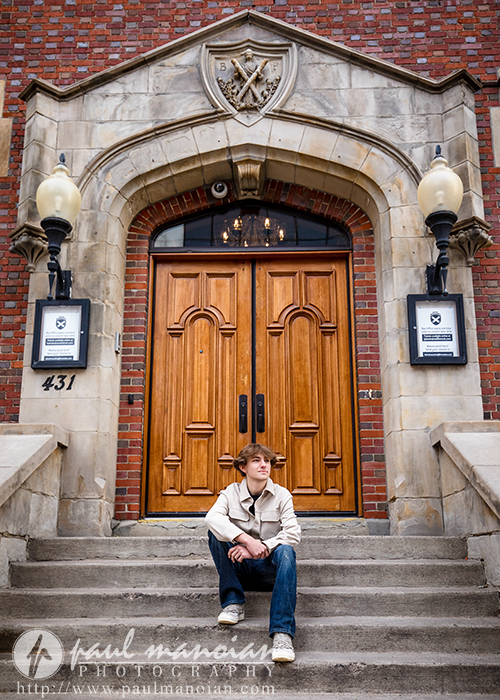 A young man in a light jacket and jeans sits on stone steps in front of large wooden double doors of a brick building with arched stone entrance and two lanterns. He looks to the side, appearing relaxed.