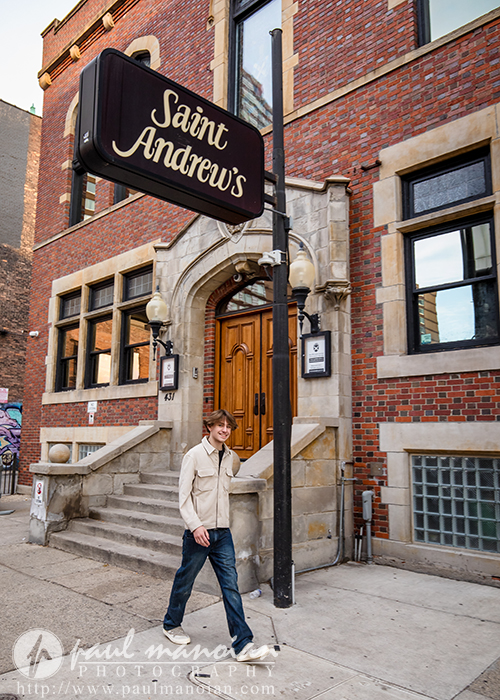 A young man walks on a sidewalk past the entrance of a brick building with a sign that reads "Saint Andrew's." The building has stone steps, arched windows, and ornate lamps by the doorway.