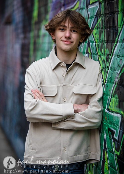 A young person with light brown hair and a beige jacket stands with arms crossed, smiling in front of a graffiti-covered brick wall during their Milford senior pictures session. The atmosphere is casual and urban.