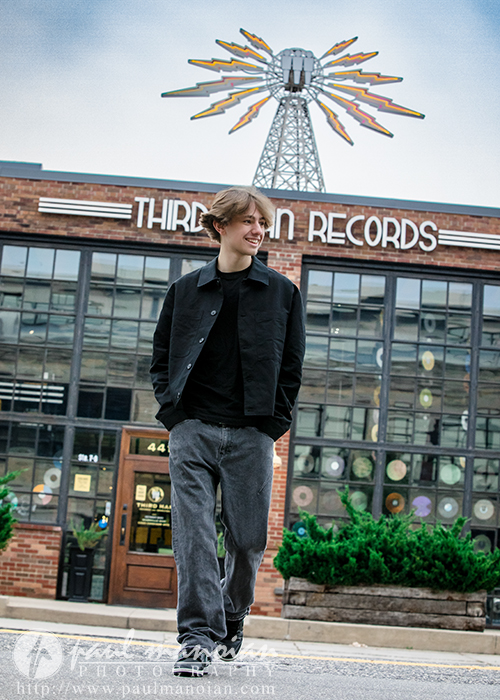 A young man with light brown hair, wearing a black jacket, black shirt, and gray jeans, smiles while walking in front of Third Man Records during his Milford senior pictures session, with a large art installation visible on the roof behind him.