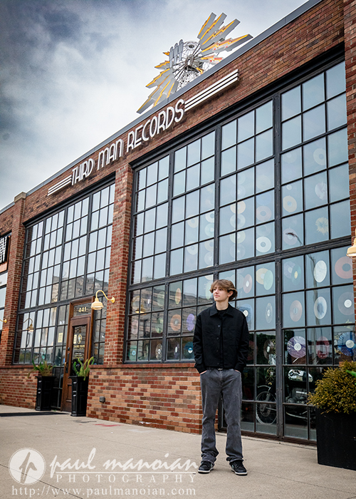 A young man stands outside Third Man Records—a brick building with large windows, vinyl records, and a silver sunburst—posing under a cloudy sky for his Milford senior pictures session.