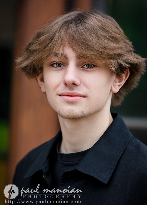 A young man with light brown, feathered hair and a short beard smiles slightly at the camera during his Milford senior pictures session, wearing a black collared shirt. The softly blurred background and photography watermark add a polished touch.