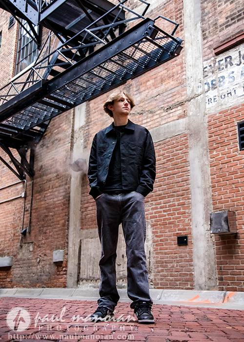 A young person with blonde hair stands in an urban alleyway, surrounded by brick walls and a metal fire escape above, looking off to the side with a relaxed posture during their Milford senior pictures session.