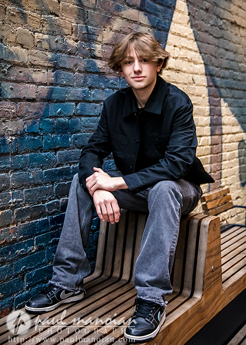 A young man with light brown hair, wearing a black jacket and gray jeans, sits on a wooden bench against a brick wall painted with a blue mural. He looks at the camera with a relaxed expression during his Milford senior pictures session.