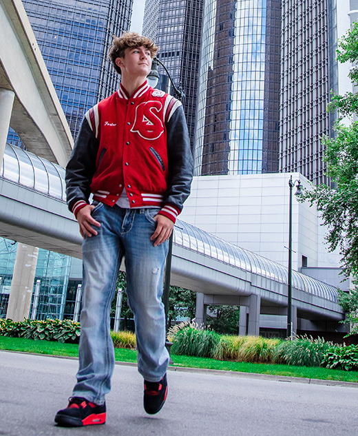 A young man in a red letterman jacket and jeans walks confidently on a city sidewalk, with tall glass buildings, greenery, and a large tree—capturing the modern vibe of his St. Clair Shores senior pictures session.