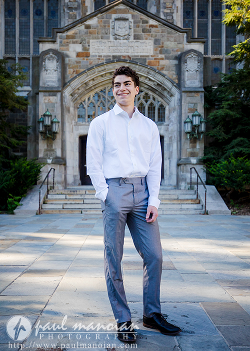 A young man wearing a white dress shirt and gray pants stands smiling in front of a large, stone university building with arched doorways and steps during his Saline senior pictures session.