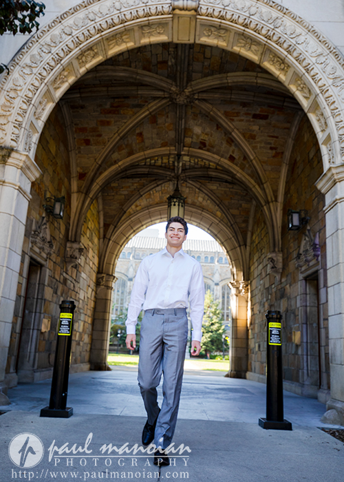 A young man in a white shirt and gray pants stands confidently under a stone archway on a university campus during his Saline senior pictures session, sunlight illuminating the entrance and historic architecture in the background.